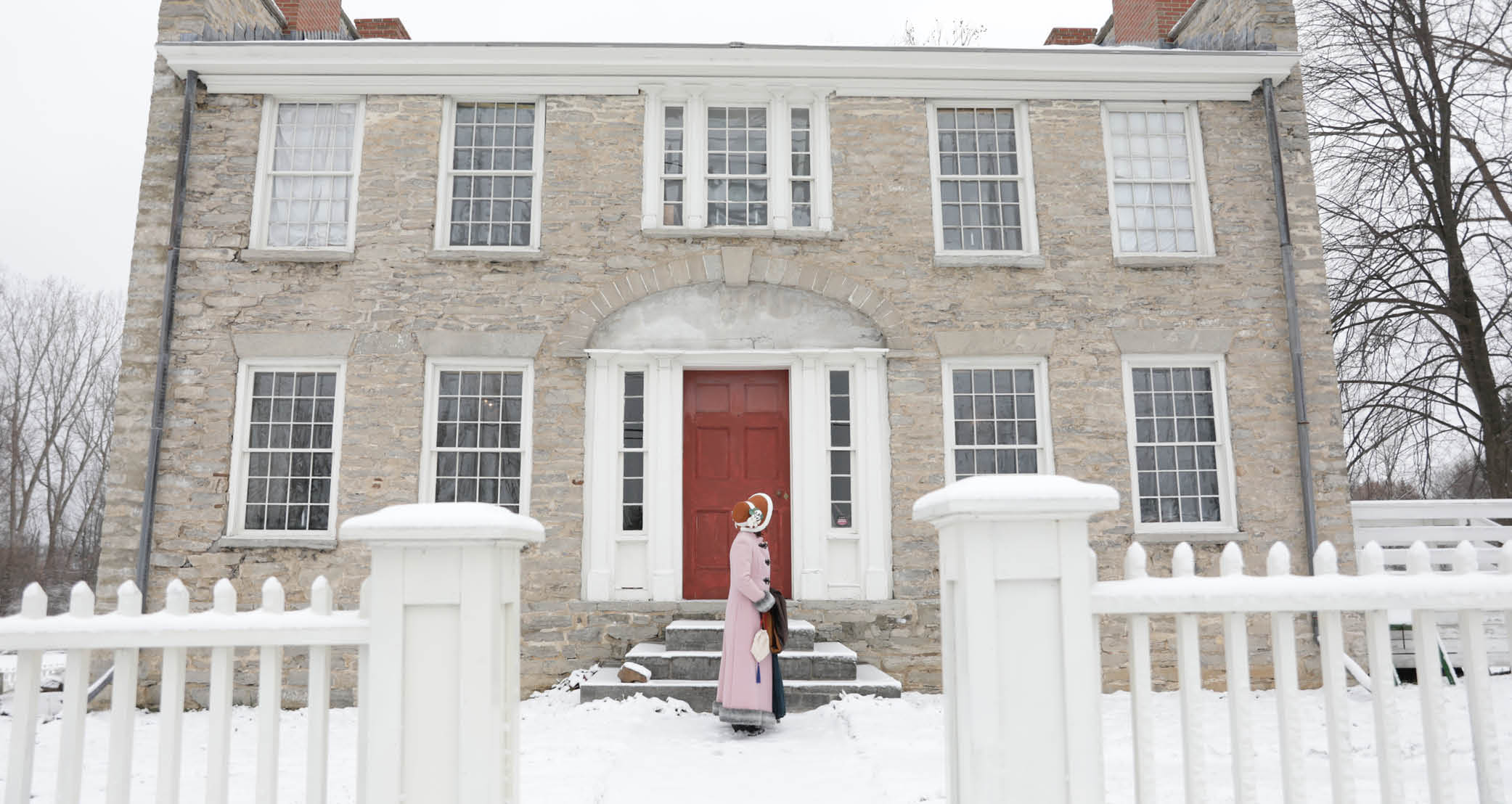 old grey stone house with a red door and a woman with a pink dress and bonnet standing on the stairs. 